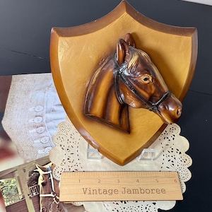 May include: A vintage horse head plaque with a shield-shaped wooden backing. The horse head is brown with a bridle, and the plaque has the text "Vintage Jamboree" on a wooden ruler. The plaque is displayed on a doily.