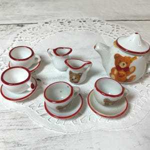 May include: A miniature white porcelain tea set with red trim. The set includes a teapot with a teddy bear design, two creamers, and four teacups with saucers. The tea set is arranged on a white doily.
