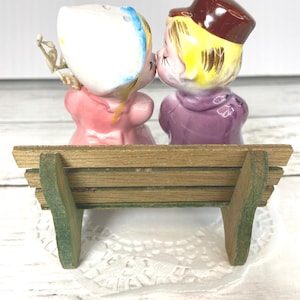 May include: Vintage ceramic salt and pepper shakers depicting a kissing couple. The figures are dressed in pink and purple clothing, with yellow hair and brown hats. They are seated on a small wooden bench with green accents. The bench is approximately 4 inches long.