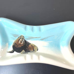May include: A ceramic ashtray with a light blue and white design. The ashtray features a walrus illustration in the center, set against a snowy landscape. The ashtray has a unique bow-tie shape.
