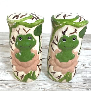 May include: A pair of ceramic salt and pepper shakers. Each shaker features a green frog design with a pink flower and green vines on a cream-colored background. The shakers are approximately 4 inches tall.