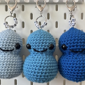 以下が含まれることがあります： 黒い目と笑顔の口を持つ、青と薄い青の毛糸で編まれたぬいぐるみが3つあります。それぞれのぬいぐるみは、単語を綴るビーズのチャームが付いた銀色のキーホルダーに付けられています。