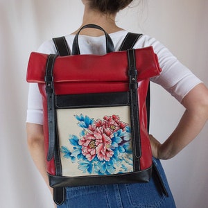 Puede incluir: Mochila de cuero roja con solapa negra y un bolsillo delantero de color crema con un diseño floral pintado a mano de peonías rosas y azules.