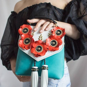 以下が含まれることがあります： 赤いポピーの花のアクセントが付いた、ターコイズと白のハンドバッグ。バッグはトップハンドル、装飾的なタッセル、金色のディテールが特徴です。花は黒い中心部を持ち、バッグの上部に配置されています。
