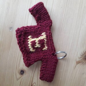 K&ouml;nnte beinhalten: Ein kleiner gestrickter Pullover mit einem gelben Buchstaben "H" auf der Vorderseite. Der Pullover ist rot und hat einen silbernen Schl&uuml;sselanh&auml;nger.