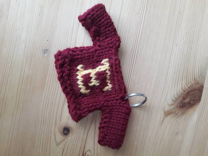 K&ouml;nnte beinhalten: Ein kleiner gestrickter Pullover mit einem gelben Buchstaben "H" auf der Vorderseite. Der Pullover ist rot und hat einen silbernen Schl&uuml;sselanh&auml;nger.