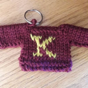 K&ouml;nnte beinhalten: Ein kleiner gestrickter, kastanienbrauner Pullover-Schl&uuml;sselanh&auml;nger mit einem aufgestickten gelben Buchstaben "K" auf der Vorderseite.