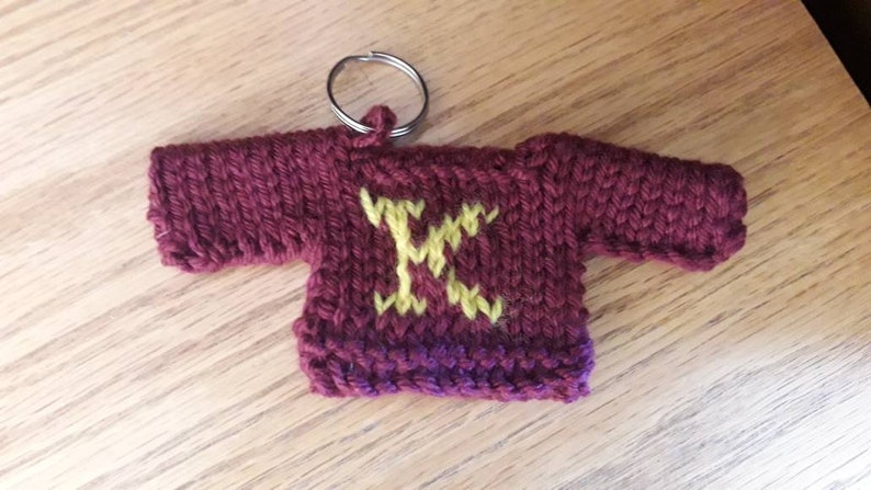 K&ouml;nnte beinhalten: Ein kleiner gestrickter, kastanienbrauner Pullover-Schl&uuml;sselanh&auml;nger mit einem aufgestickten gelben Buchstaben "K" auf der Vorderseite.