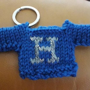 K&ouml;nnte beinhalten: Ein blauer gestrickter Pullover-Schl&uuml;sselanh&auml;nger mit einem silbernen Buchstaben H auf der Vorderseite. Der Schl&uuml;sselanh&auml;nger ist an einem silbernen Schl&uuml;sselring befestigt.