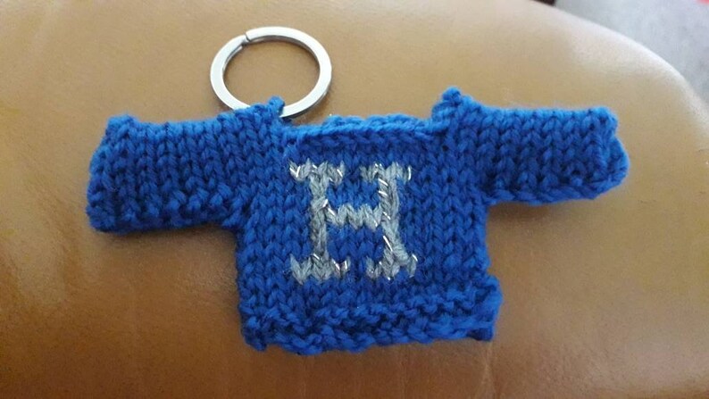 K&ouml;nnte beinhalten: Ein blauer gestrickter Pullover-Schl&uuml;sselanh&auml;nger mit einem silbernen Buchstaben H auf der Vorderseite. Der Schl&uuml;sselanh&auml;nger ist an einem silbernen Schl&uuml;sselring befestigt.