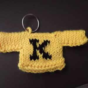 K&ouml;nnte beinhalten: Ein gelber gestrickter Pullover-Schl&uuml;sselanh&auml;nger mit einem schwarzen Buchstaben "M" auf der Vorderseite.