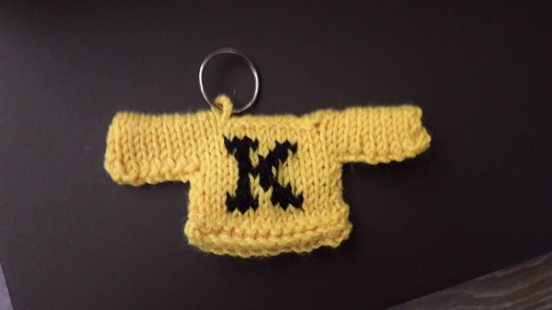 K&ouml;nnte beinhalten: Ein gelber gestrickter Pullover-Schl&uuml;sselanh&auml;nger mit einem schwarzen Buchstaben "M" auf der Vorderseite.