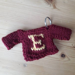 K&ouml;nnte beinhalten: Ein kleiner gestrickter Pullover-Schl&uuml;sselanh&auml;nger mit rotem K&ouml;rper und gelbem Buchstaben E auf der Vorderseite. Der Schl&uuml;sselanh&auml;nger hat einen silbernen Ring, der oben befestigt ist.