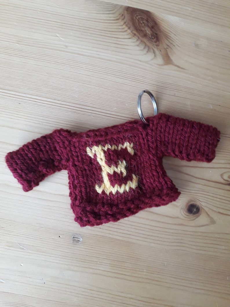 K&ouml;nnte beinhalten: Ein kleiner gestrickter Pullover-Schl&uuml;sselanh&auml;nger mit rotem K&ouml;rper und gelbem Buchstaben E auf der Vorderseite. Der Schl&uuml;sselanh&auml;nger hat einen silbernen Ring, der oben befestigt ist.