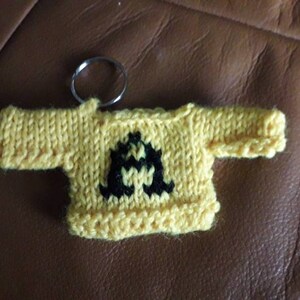 K&ouml;nnte beinhalten: Ein gelber gestrickter Pullover-Schl&uuml;sselanh&auml;nger mit einem schwarzen Buchstaben A auf der Vorderseite.