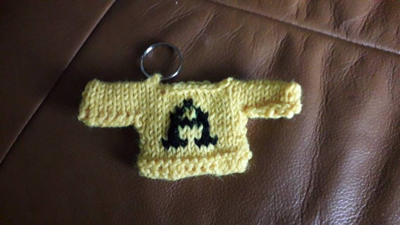 K&ouml;nnte beinhalten: Ein gelber gestrickter Pullover-Schl&uuml;sselanh&auml;nger mit einem schwarzen Buchstaben A auf der Vorderseite.