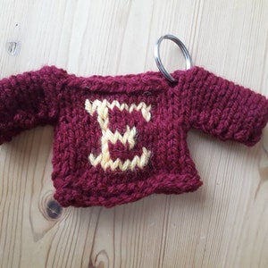 K&ouml;nnte beinhalten: Ein kleiner, gestrickter burgunderfarbener Pullover mit einem gelben Buchstaben "E" auf der Vorderseite. Der Pullover ist an einem silbernen Schl&uuml;sselring befestigt.
