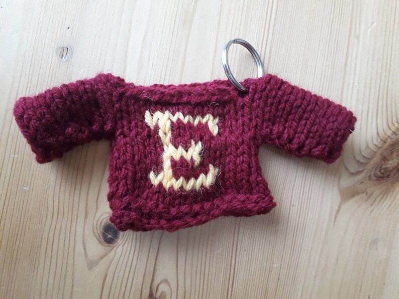 K&ouml;nnte beinhalten: Ein kleiner, gestrickter burgunderfarbener Pullover mit einem gelben Buchstaben "E" auf der Vorderseite. Der Pullover ist an einem silbernen Schl&uuml;sselring befestigt.