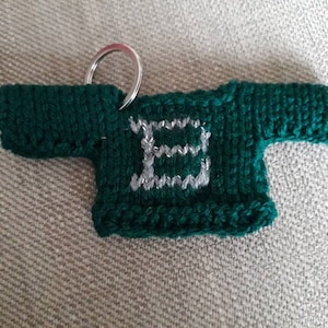 K&ouml;nnte beinhalten: Ein gr&uuml;ner gestrickter Pullover-Schl&uuml;sselanh&auml;nger mit einem silbernen Buchstaben "E" auf der Vorderseite.