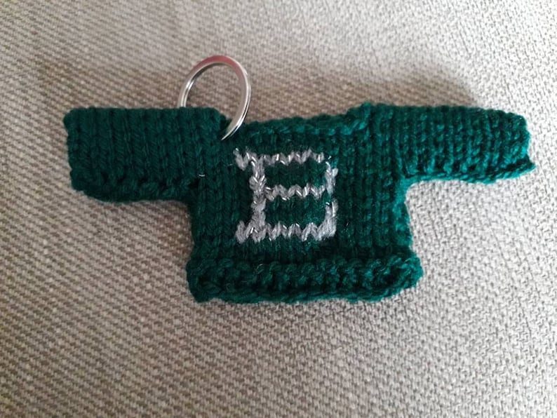 K&ouml;nnte beinhalten: Ein gr&uuml;ner gestrickter Pullover-Schl&uuml;sselanh&auml;nger mit einem silbernen Buchstaben "E" auf der Vorderseite.