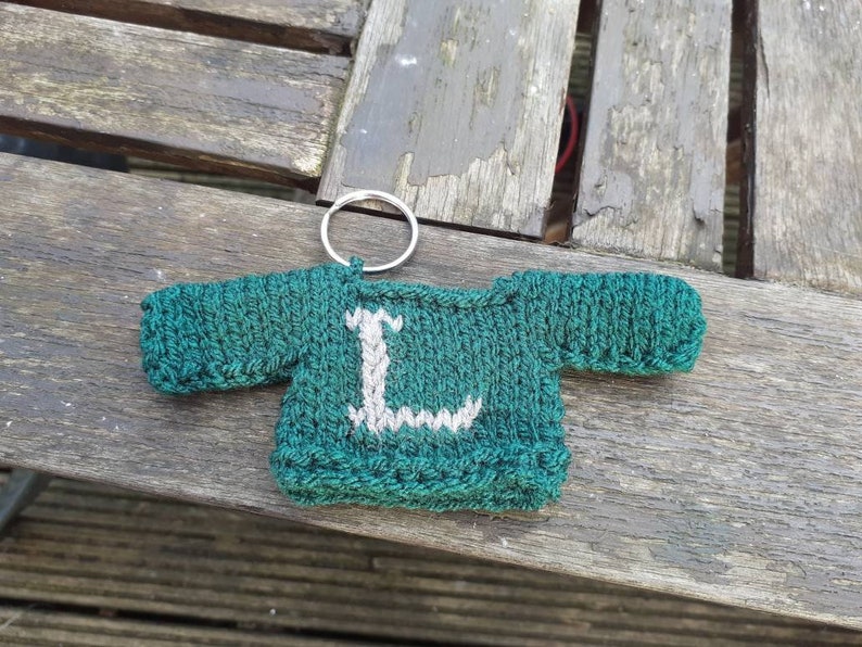 K&ouml;nnte beinhalten: Ein gr&uuml;ner gestrickter Pullover-Schl&uuml;sselanh&auml;nger mit einem wei&szlig;en Buchstaben L auf der Vorderseite. Der Schl&uuml;sselanh&auml;nger ist an einem silbernen Schl&uuml;sselring befestigt.