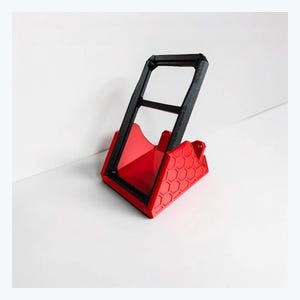 May include: A red plastic stand with a textured hexagonal pattern on the sides and a black rectangular frame attached at an angle. The stand features a hollow center and a sturdy base designed to hold an object in an upright, tilted position.