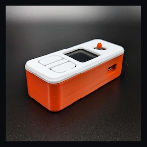 May include: A rectangular electronic device with a bright orange base and a white top panel. The top features two white rectangular buttons, a small black display screen, and a single protruding orange circular button on the right side.