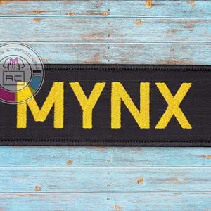 May include: A rectangular black patch with the word "MYNX" embroidered in yellow thread. The patch has a black border and is set against a blue and brown wooden background. The patch is likely a military or tactical patch.