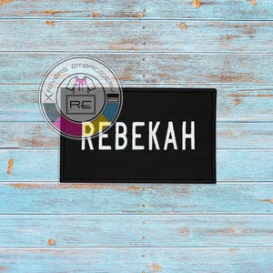 May include: Black patch with white text that reads "REBEKAH" on a light blue wood background.