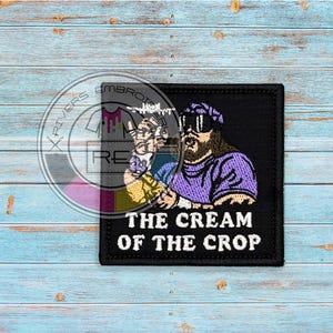 Peut inclure: Écusson carré noir avec une image brodée d'un homme portant des lunettes de soleil et un haut violet. Le texte "THE CREAM OF THE CROP" est imprimé en blanc. Fond en bois bleu.
