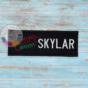May include: A rectangular black embroidered patch featuring the white text SKYLAR in capital letters. To the left of the name, the text HO HO HO is embroidered in red, and the word WOOF! is embroidered in green, both in a stylized font.