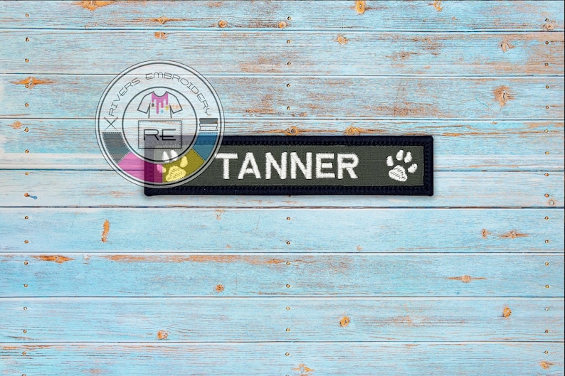May include: A black and green dog tag with white text that reads "TANNER" and a white paw print on the right side.