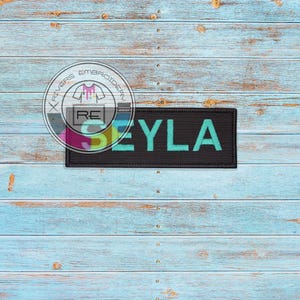 Pode incluir: Etiqueta de tecido preto com o nome "SEYLA" bordado em letras verde turquesa.