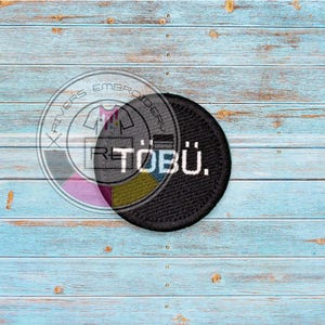 Könnte beinhalten: Schwarzer gestickter Aufnäher mit weißem Text, der "RETOBÜ." lautet. Der Aufnäher befindet sich auf einem hellblauen Holzhintergrund.