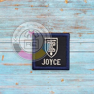 May include: A navy blue embroidered patch with the name "JOYCE" in white block letters. The patch features a white and blue crest design on a black background, with a navy blue border. The patch is square and sits on a light blue wooden surface.