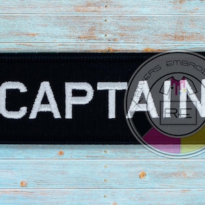 May include: Black embroidered patch with white text that reads "CAPTAIN". The patch has a circular logo in the bottom right corner with the text "Embroidery" and a stylized image of a shirt.