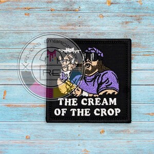 May include: Black embroidered patch with a cartoon image of a man with a beard and sunglasses wearing a purple shirt. The text "The Cream of the Crop" is below the image.