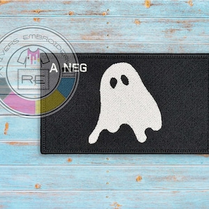 Op de afbeelding: Zwart rechthoekig embleem met een wit spookontwerp en twee ovale ogen. De patch heeft een gestikte rand en de tekst "A NEG" is zichtbaar. Geschikt voor kleding of accessoires.