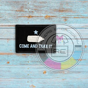 May include: Black embroidered patch with a white star and a white silhouette of a ship. The text "Come and Take It" is embroidered in white below the ship. The patch is on a blue wooden background.