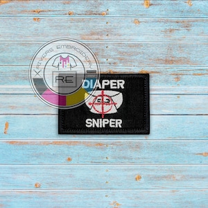 May include: Black embroidered patch with white text that reads "Diaper Sniper" and a white diaper with a red target symbol.