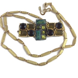 Puede incluir: Un collar de cadena dorada con un colgante geométrico con piedras negras, verdes y amarillas.