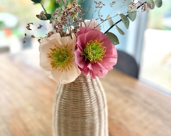 Op de afbeelding: Een gevlochten vaas met een bloemstuk. Het arrangement bevat twee grote klaproosachtige bloemen, één licht perzik en één roze, met gele en groene centra. Eucalyptus en kleine, gedroogde bloemen maken het geheel compleet. De vaas is beige en heeft een getextureerd, geweven uiterlijk.