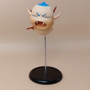 May include: A cartoon figurine of a creature with a light tan body, blue hair, and angry blue eyebrows. It has small horns and is mounted on a silver rod with a black circular base. The creature's mouth is open, revealing teeth.