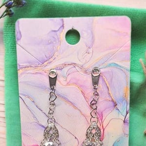 Puede incluir: Pendientes colgantes plateados con una gran gema redonda y transparente. Los pendientes se exhiben en una tarjeta decorativa con un diseño veteado en rosa, morado y dorado, sobre un fondo verde.