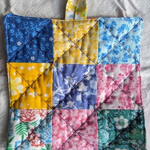 Puede incluir: Un agarrador de cocina cuadrado acolchado con una presilla para colgar. Está hecho de nueve cuadrados de tela con varios estampados florales y colores, incluyendo azul, amarillo, rosa y verde. Las costuras negras delinean los cuadrados.