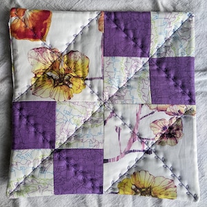 Puede incluir: Un agarrador de olla de tela acolchada blanca con un diseño de patchwork. La tela es una mezcla de patrones blancos, morados y florales. El agarrador de olla está cosido con una puntada decorativa.