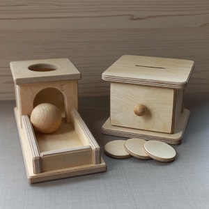 May include: Two wooden toy boxes. One box has a drawer and a knob. The other box has a hole in the top and a ramp with a wooden ball on it. There are four wooden circles in front of the boxes.
