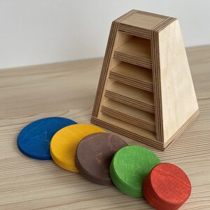 May include: Wooden stacking toy with five colorful circles. The circles are blue, yellow, brown, green, and red. The toy is designed to help children learn about colors and shapes.