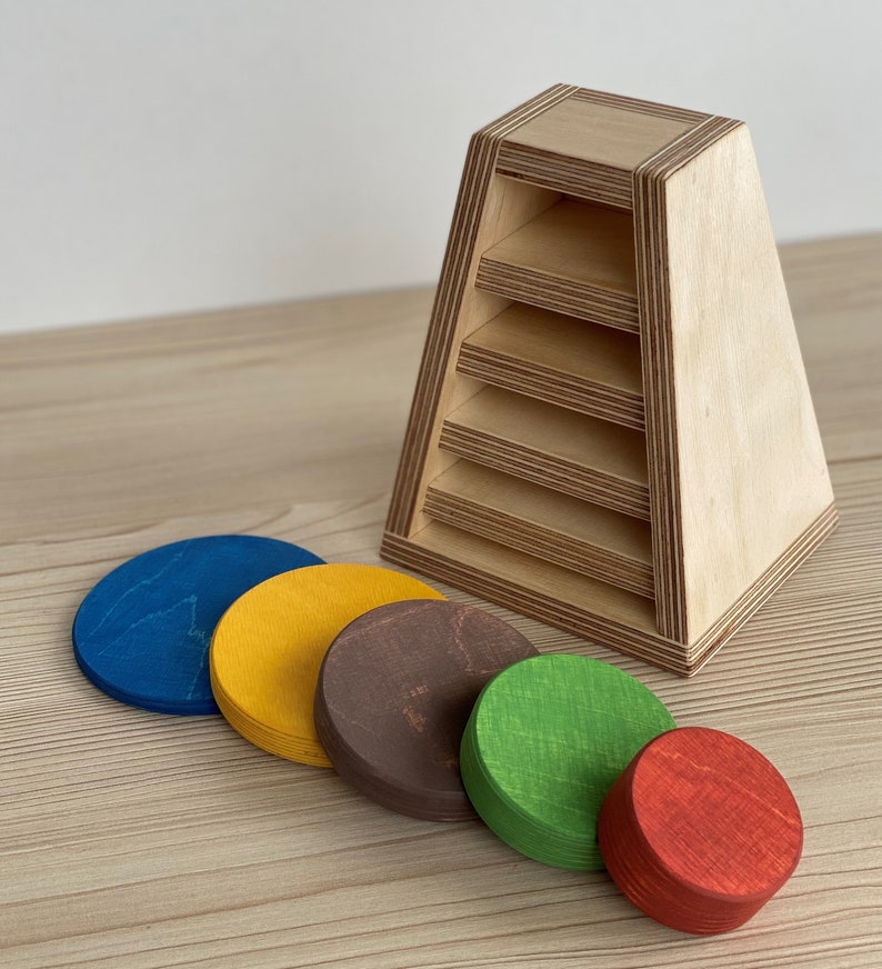 May include: Wooden stacking toy with five colorful circles. The circles are blue, yellow, brown, green, and red. The toy is designed to help children learn about colors and shapes.