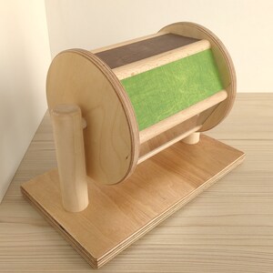May include: A wooden toy with a spinning wheel and a green fabric panel. The wheel is mounted on a wooden base with a wooden post.